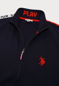 Marinblå zip-up jacka med röd "PLAY" text på kragen, röd logobroderi och flerfärgad randig dragkedjedetalj. Mjuk tyg.
