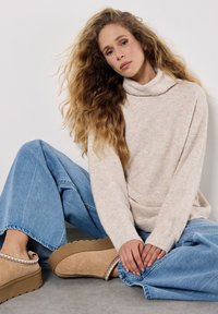 Beżowy sweter z golfem o miękkiej teksturze, zestawiony z jasnoniebieskimi szerokimi spodniami dżinsowymi. Brązowe zamszowe buty wsuwane, z dekoracyjnym wykończeniem.