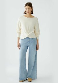 Cremiger gestrickter Pullover mit gedrehtem Vorderteil, kombiniert mit hellblauen weiten Jeans und beigefarbenen Slipschuhen, vor einem einfarbigen Hintergrund.