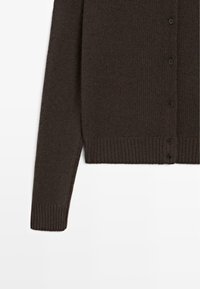 Mørkebrun strikket cardigan med knaplukning, ribbede manchetter og kant, vist fra torsoen til en ærme på en hvid baggrund.