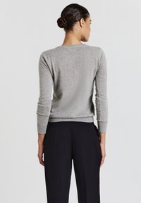 Donna con i capelli scuri raccolti in un bun, che indossa un maglione a maniche lunghe grigio chiaro e pantaloni neri, vista di schiena su uno sfondo neutro.