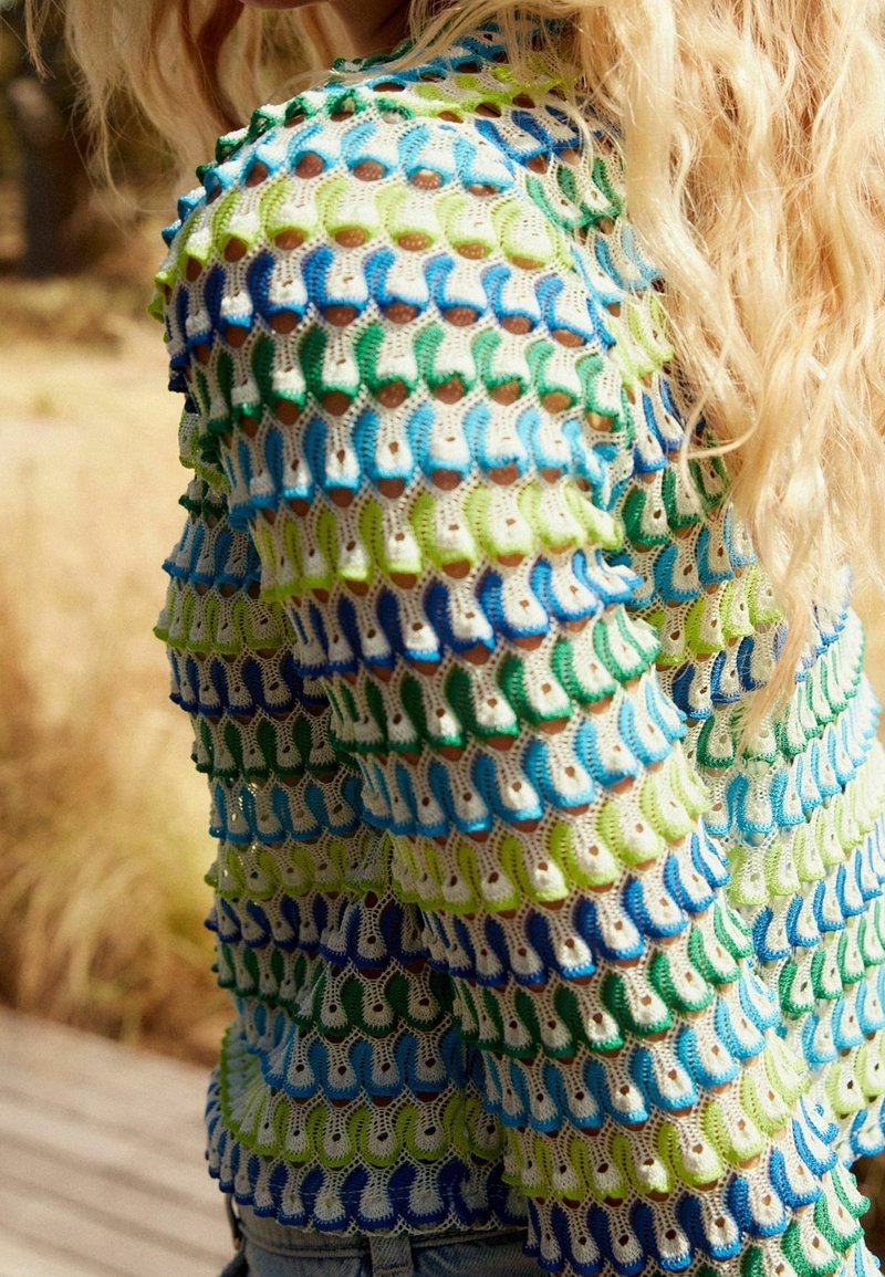 Femme aux longs cheveux blonds portant un pull ajouré multicolore en bleu, vert et blanc, debout à l'extérieur près de l'herbe sèche.