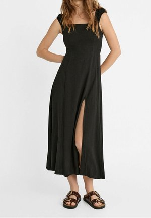 Femme portant une robe midi noire sans manches avec une fente frontale, assortie à des sandales marron foncé ornées de grosses boucles et de clous.