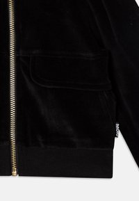 Giacca in velluto nero con zip dorata, polsini a costine e due tasche frontali. Presenta un'etichetta con logo sul lato. Texture morbida.