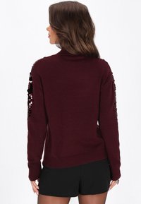 Burgundowy sweter o dopasowanym kroju, z cekinowymi akcentami na rękawach, prążkowanymi mankietami i dołem. Miękka tekstura, casualowy styl.