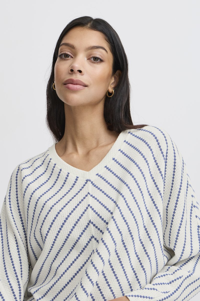 Frau mit langen dunklen Haaren trägt einen weißen Pullover mit blauen diagonalen Streifen und goldenen Creolen, posiert vor einem schlichten hellen Hintergrund.