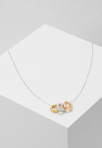 Collier en chaîne argentée avec trois anneaux entrelacés en or, argent avec diamants, et or rose sur une surface géométrique blanche.