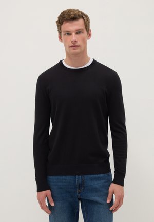 Hombre con cabello castaño claro, vestido con un suéter negro de manga larga sobre una camisa blanca y jeans azules, de pie frente a un fondo liso y claro.