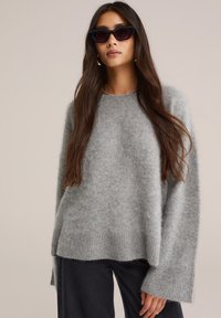 Jasnoszary, oversize sweter z prążkowanym dołem i mankietami, wykonany z miękkiego, puszystego materiału. Modelka nosi czarne okulary przeciwsłoneczne oraz dżinsy.