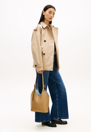 Γυναίκα που φοράει μπεζ διπλόμπουτο σακάκι, μπλε τζιν παντελόνι με φαρδιά πόδια, μαύρα μοκασίνια, κρατώντας μια καφέ δερμάτινη τσάντα με ανοιχτό μπλε φόδρα.