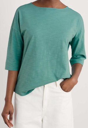 Personne portant un haut ample à manches trois-quarts de couleur sarcelle légèrement rentré dans un pantalon blanc taille haute, main posée dans la poche.