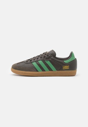 Tummanruskea Adidas Samba -lenkkari vihreillä raidoilla, vihreällä kantalenkillä, kuminvärisellä pohjalla ja kultaisella "adidas SAMBA" -logolla sivussa.