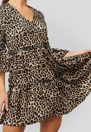 Frau trägt ein braun-schwarzes Leopardenmuster V-Ausschnitt Kleid mit Ellbogen-langen Rüschensäumen und hält den Rock seitlich ausgestreckt.