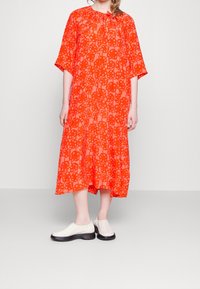 Robe orange jusqu'au genou avec un motif floral, manches courtes et encolure froncée. Associée à des chaussures blanches sans lacets.