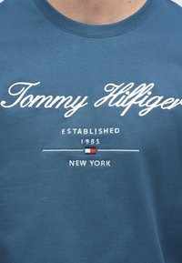 Μπλε βαμβακερό t-shirt με λευκό κεντημένο κείμενο "Tommy Hilfiger", με επιπλέον κείμενο "ESTABLISHED 1985 NEW YORK" και διακοσμητικό λογότυπο.