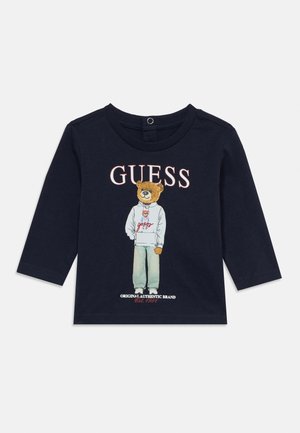 Maglietta blu navy a maniche lunghe per bambino con orsetto che indossa una felpa con cappuccio e jeans, logo "GUESS" sopra e scritta del marchio sotto.