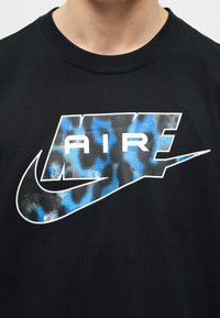 Tricou negru cu un logo mare „AIR” în model camuflaj albastru și gri și cu swoosh-ul Nike pe piept.