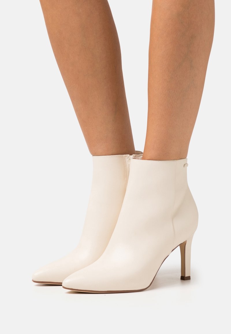 Jambes portant des bottines à bout pointu blanc cassé avec des talons fins de hauteur moyenne et des fermetures éclair latérales sur un fond blanc.