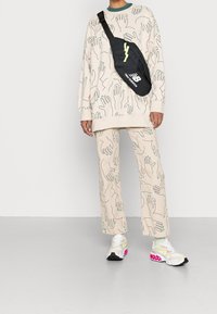 Persoon in een beige bijpassende set met abstracte handafdrukken, witte sneakers met roze accenten en een zwarte New Balance schoudertas.