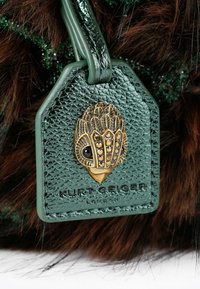 Etichetta in pelle verde con texture scolpita e motivo animale dorato, attaccata a un accessorio in pelliccia marrone. I dettagli mostrati includono il nome del marchio.
