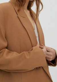Blazer beige avec un revers cranté, texture lisse, coupe décontractée et détails de couture subtils. Manches longues avec boutons en accent.