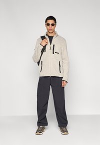 Μπεζ φλις μπουφάν με μαύρα φερμουάρ και λεπτομέρειες, σε συνδυασμό με σκούρο παντελόνι cargo και γκρι αθλητικά παπούτσια. Το μοντέλο φορά γυαλιά ηλίου και κρατά σακίδιο πλάτης.