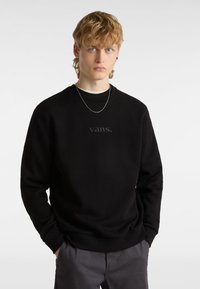 Sudadera negra con un cuello redondo y puños acanalados, que presenta un logo "vans." bordado en tono sobre el pecho. Combinada con pantalones gris oscuro.