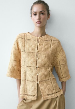 Giovane donna che indossa un cardigan beige lavorato a uncinetto con bottoni, motivi floreali e bordi smerlati, in piedi davanti a uno sfondo chiaro.