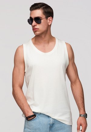 Jeune homme portant des lunettes de soleil noires, un débardeur blanc, un jean bleu clair, une montre et des bracelets noirs, posant avec une main dans la poche.