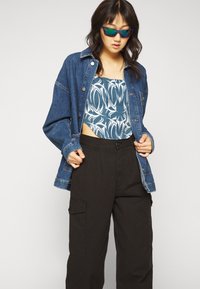 Farkkutakki napin kiinnityksellä, sinivalkoinen kuvioitu crop top, mustat cargo-housut taskuilla ja sinisävyiset aurinkolasit.