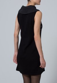 Kvinna i en ärmlös svart klänning med texturerad vikkrage, sedd bakifrån mot en enfärgad grå bakgrund.