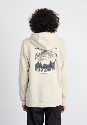 Persoon draagt een beige hoodie met een afbeelding van een berg en bos en de tekst "Columbia Portland, Oregon" op de achterkant, kijkt naar achteren.