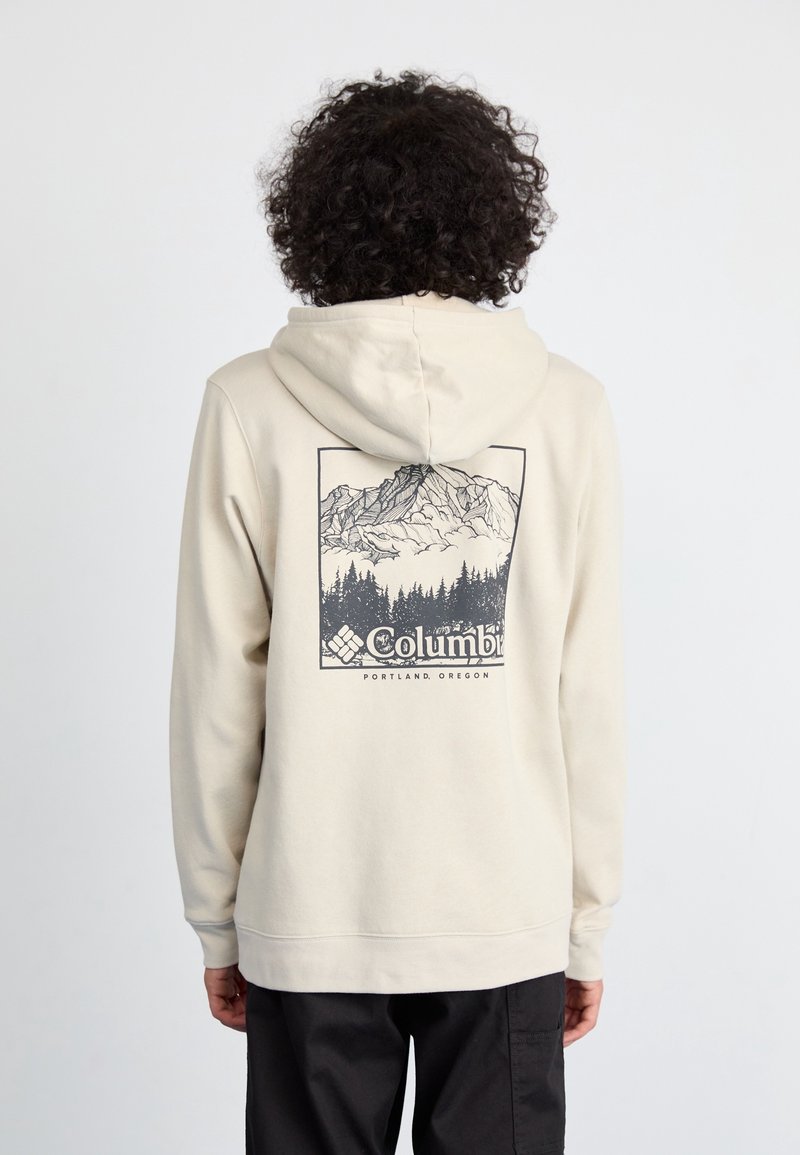 Personne portant un sweat à capuche beige avec un motif de montagne et de forêt, ainsi que le texte « Columbia Portland, Oregon » dans le dos, vue de dos.