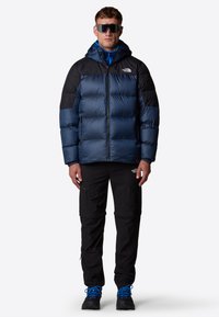 Kurtka puchowa w granatowym kolorze z czarnymi akcentami, z zapięciem na zamek, kapturem i pikowanym wzorem. Połączona z czarnymi spodniami cargo i niebieskimi butami do wędrówek.