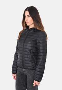 Donna con capelli lunghi che indossa una giacca nera trapuntata con zip e jeans grigi, in piedi contro uno sfondo chiaro e uniforme.