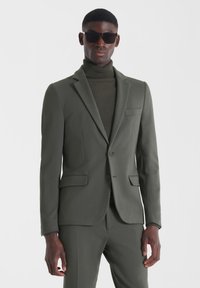 Traje verde oliva con un blazer ajustado y pantalones a juego. Cuenta con un bolsillo de pecho y cierre de dos botones. Suéter de cuello alto texturizado por debajo.