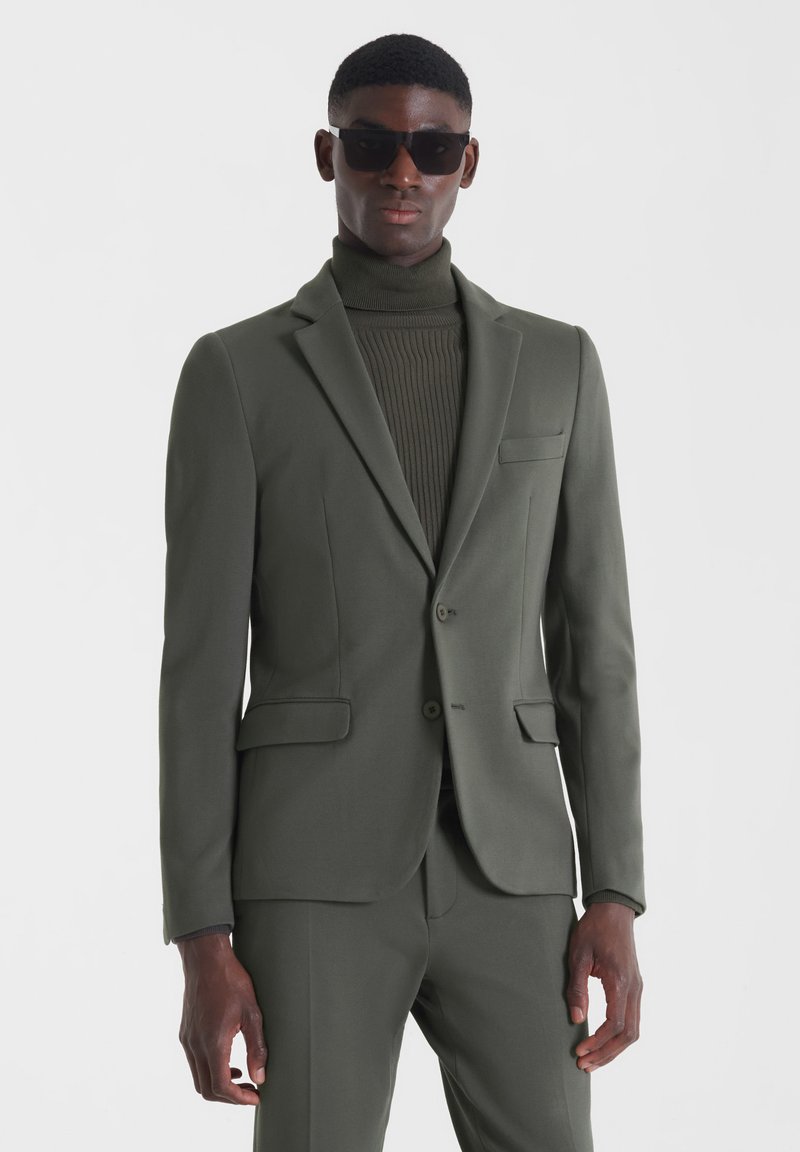 Traje verde oliva con un blazer ajustado y pantalones a juego. Cuenta con un bolsillo de pecho y cierre de dos botones. Suéter de cuello alto texturizado por debajo.