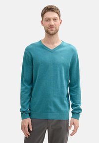 Teal V-Ausschnitt-Pullover aus weichem Stoff, mit langen Ärmeln, geripptem Saum und lässiger Passform, vor einem schlichten weißen Hintergrund präsentiert.