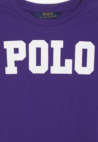 Paarse crewneck shirt met grote witte blokletters die "POLO" spellen en een klein marineblauw label met gouden tekst "POLO RALPH LAUREN" binnenin de kraag.