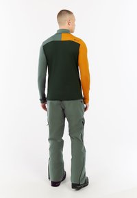 Fleece pullover in groen en oranje kleurblokken met hoge kraag; gecombineerd met lichtgroene broek met ritsen bij de enkels.