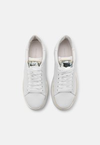 Sneaker bianchi in pelle con una texture liscia, caratterizzati da dettagli dorati e una gemma decorativa sulla linguetta. Design classico con lacci.