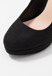 Gros plan sur l'avant d'une chaussure à talon haut en daim noir avec un bout arrondi et une doublure intérieure beige.