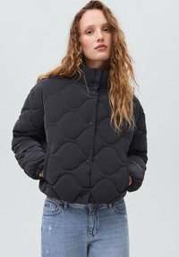 Veste noire matelassée avec un col haut, présentant des motifs arrondis et une fermeture à boutons-pression, associée à un jean en dégradé bleu clair.