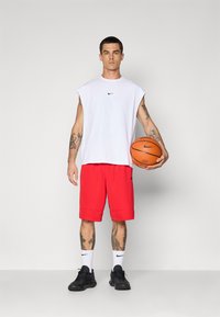 Man with tattoos wearing sleeveless white shirt, red shorts, white Nike socks, black shoes, holding an orange basketball against gray background.