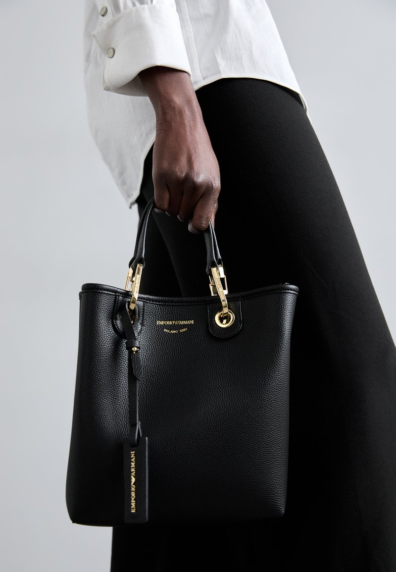Borsa a mano in pelle nera con finitura testurizzata, hardware dorato, dettaglio del logo e un'etichetta removibile. Due manici corti e forma strutturata.