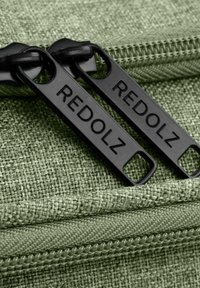 Due cerniere nere con etichetta "REDOLZ" su un tessuto intrecciato verde con una traccia di cerniera visibile.