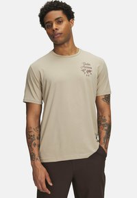 Beige t-shirt met korte mouwen, voorzien van een logo-print aan de voorkant, een ronde halslijn en een zachte textuur. Draagt het met donkere broek, waarop gebronzeerde tatoeages te zien zijn.