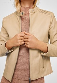 Žena v béžové kožené bundě s prošívanými detaily na ramenech, má na sobě pletený svetr v narůžovělé hnědé barvě, ruce má sepnuté před sebou, obličej není vidět.
