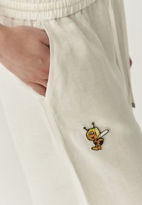 Licht beige broek van een textuurstof. Bevat een zak en een geborduurde cartoonbij aan de zijkant. Elastische tailleband.