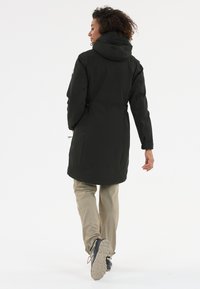 Zwarte, knielange jas met een capuchon, verstelbare taillebanden en lange mouwen. Draag het over lichtbeige broeken en donkere schoenen.